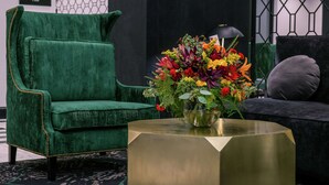 A green velvet armchair, a gold side table with a floral arrangement, and a grey sofa in a modern interior setting.