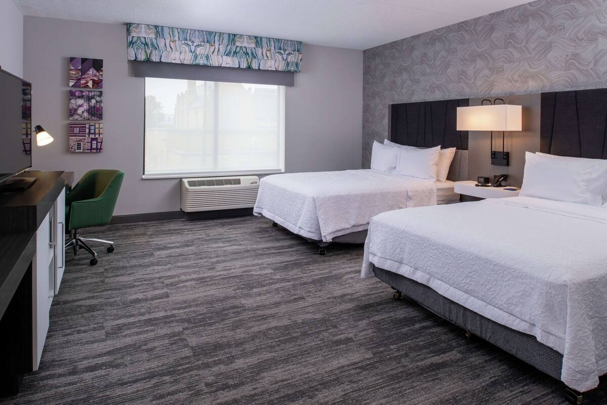 A hotel room with two beds, a desk, a chair, and a large window.