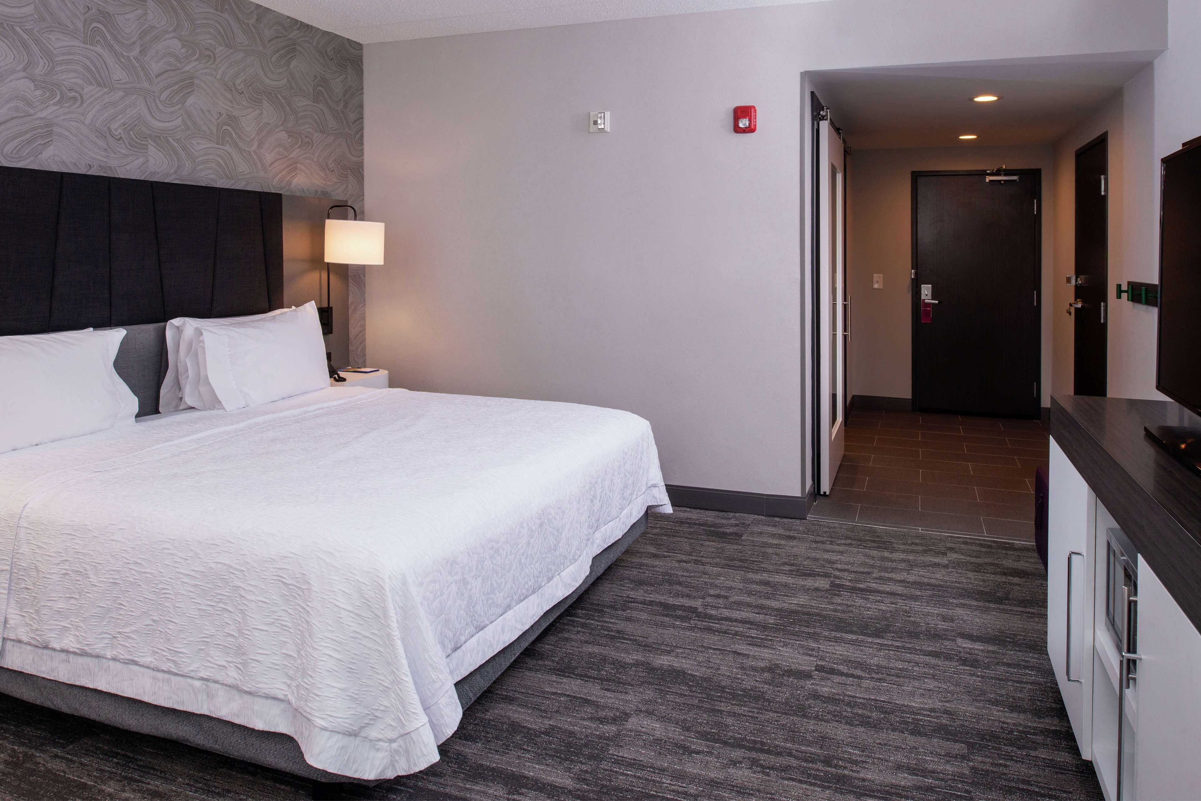 A hotel room with a large bed, a nightstand, a lamp, a flat-screen TV, and a door leading to a hallway.