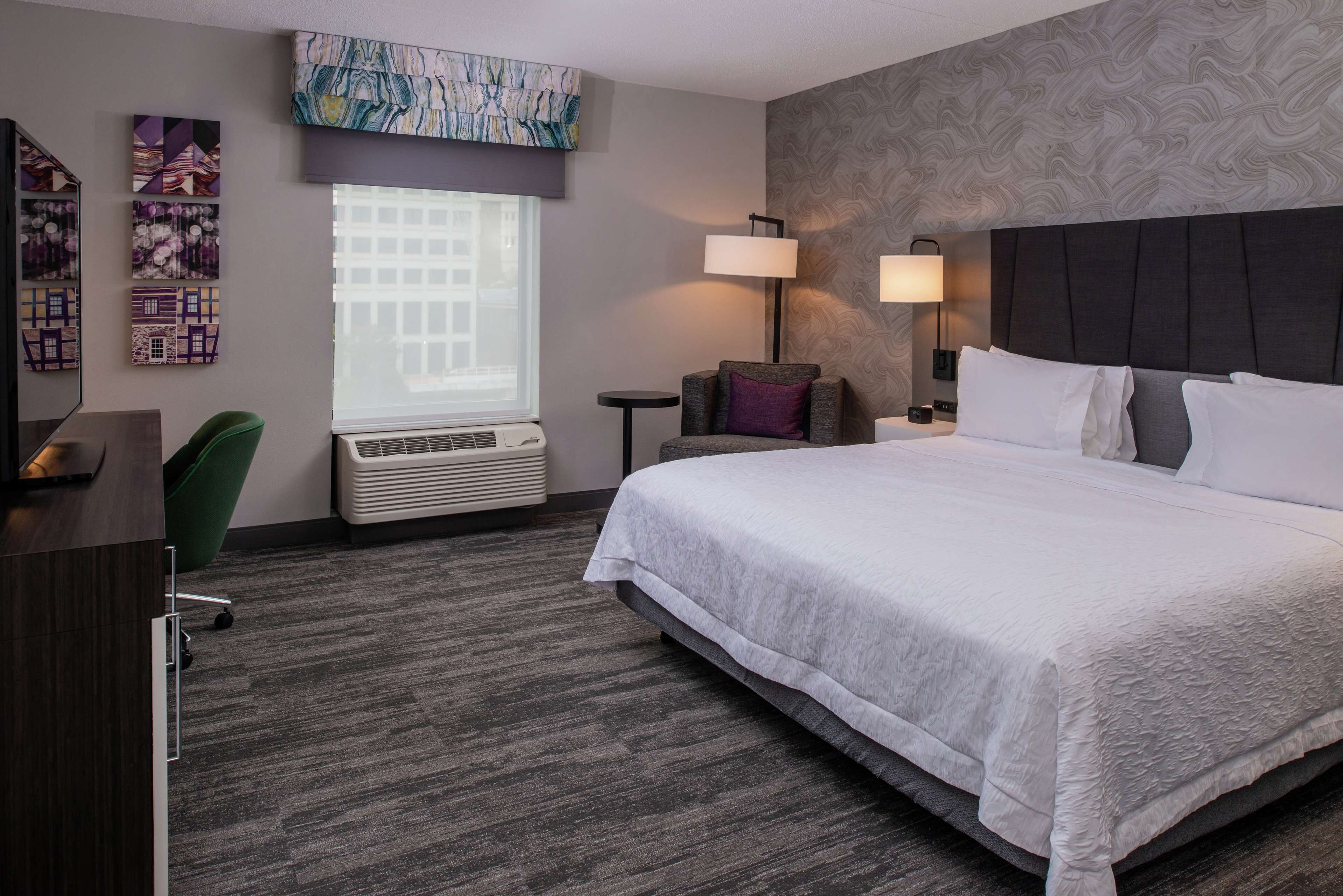 Habitación de hotel con una cama grande, un escritorio, una silla, un ventanal con persianas, un aire acondicionado de pared y dos lámparas de pared.