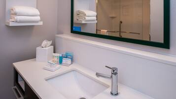 A hotel bathroom with a white sink, a mirror with a green frame, and shelves with folded towels.