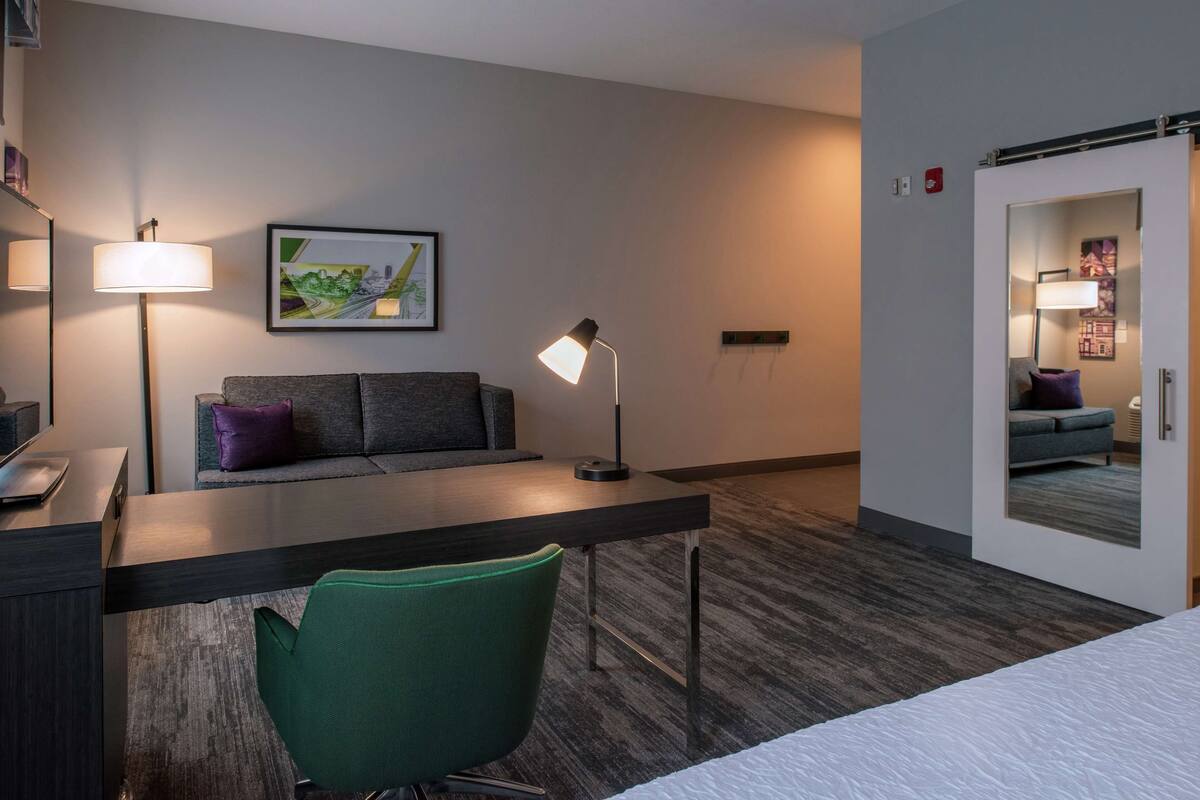 A hotel room with a desk, a green chair, a sofa with purple cushions, a standing lamp, and a framed picture on the wall.