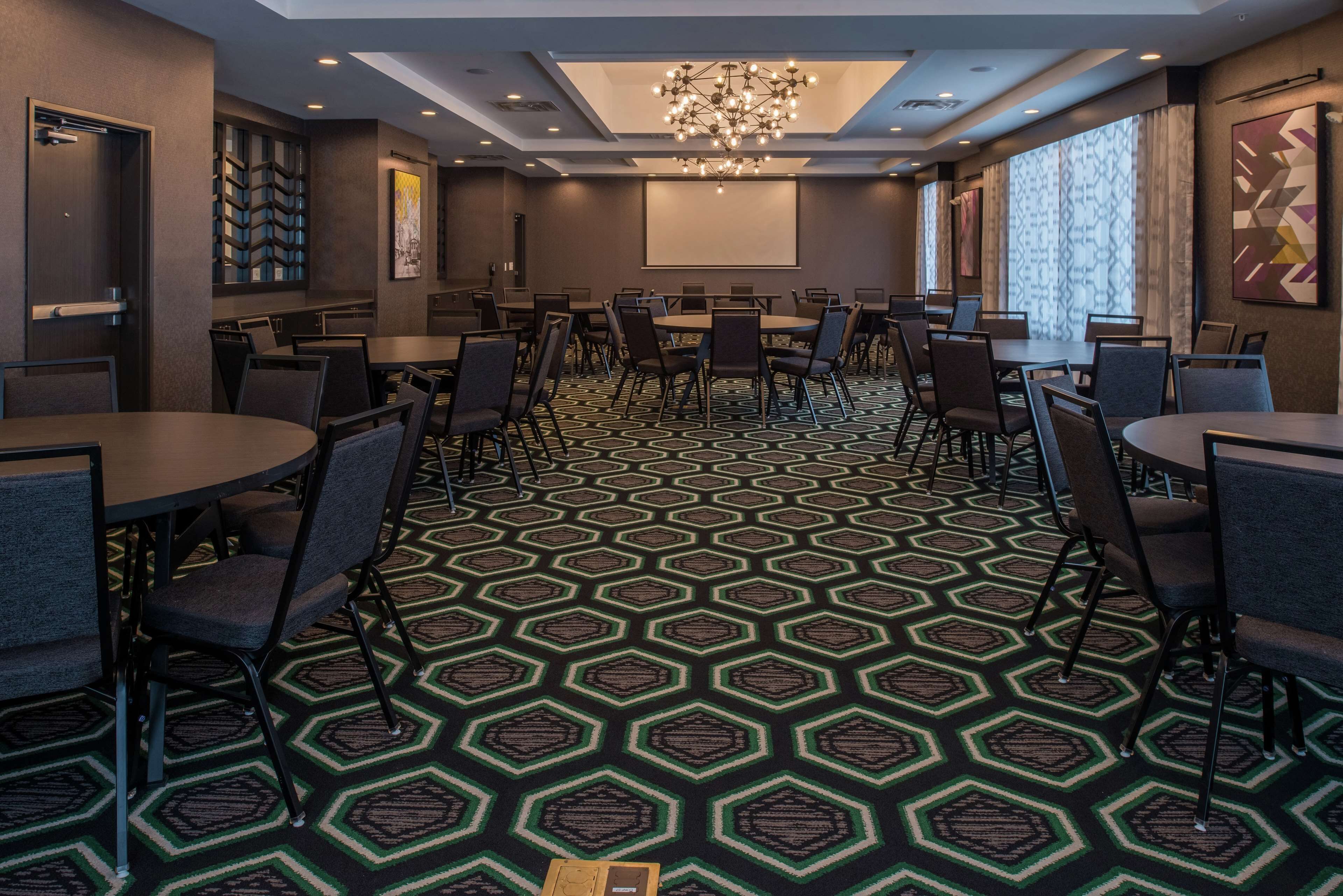 A conference room with multiple tables and chairs, a patterned carpet, a chandelier, and a projector screen on the wall.