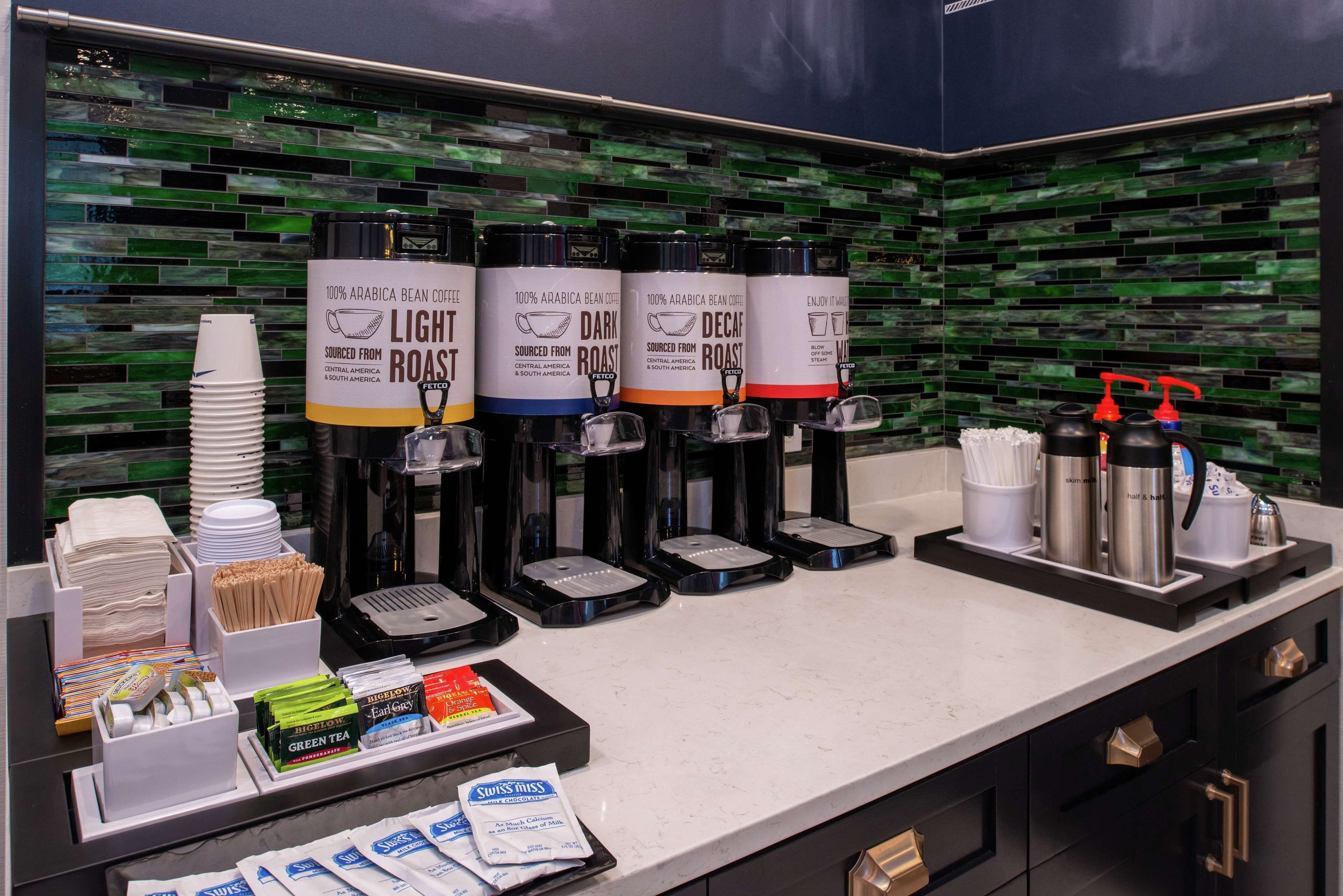 A coffee station with four coffee dispensers labeled 'Light Roast,' 'Dark Roast,' 'Decaf,' and 'Enjoy It.' There are also coffee cups, a coffee pot, and a tray with coffee mugs and a thermos.