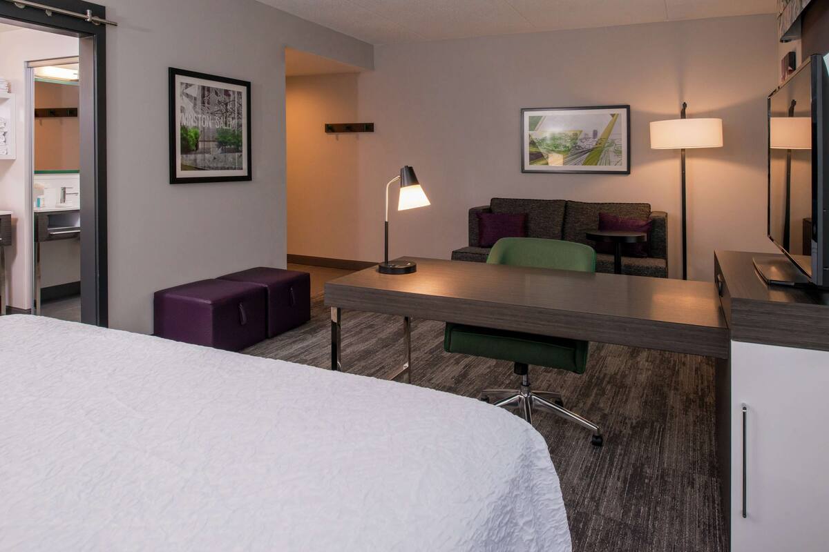 A hotel room with a bed, a desk, a chair, a sofa, a purple ottoman, and a lamp.