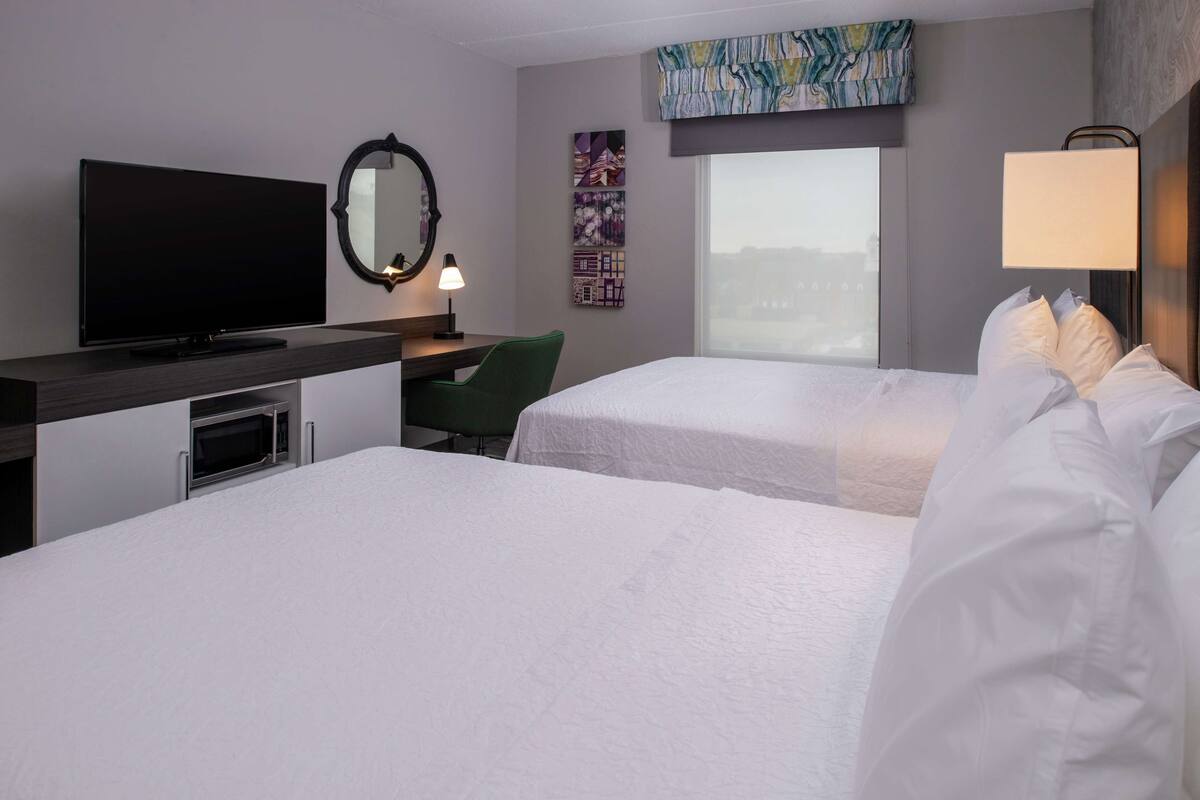 A hotel room with a flat-screen TV, a desk with a lamp, two beds with white linens, a green chair, and artwork on the walls.