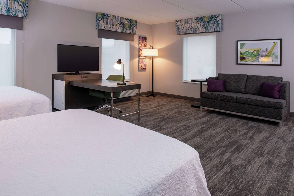 A hotel room with two beds, a desk with a TV, a lamp, and a couch.