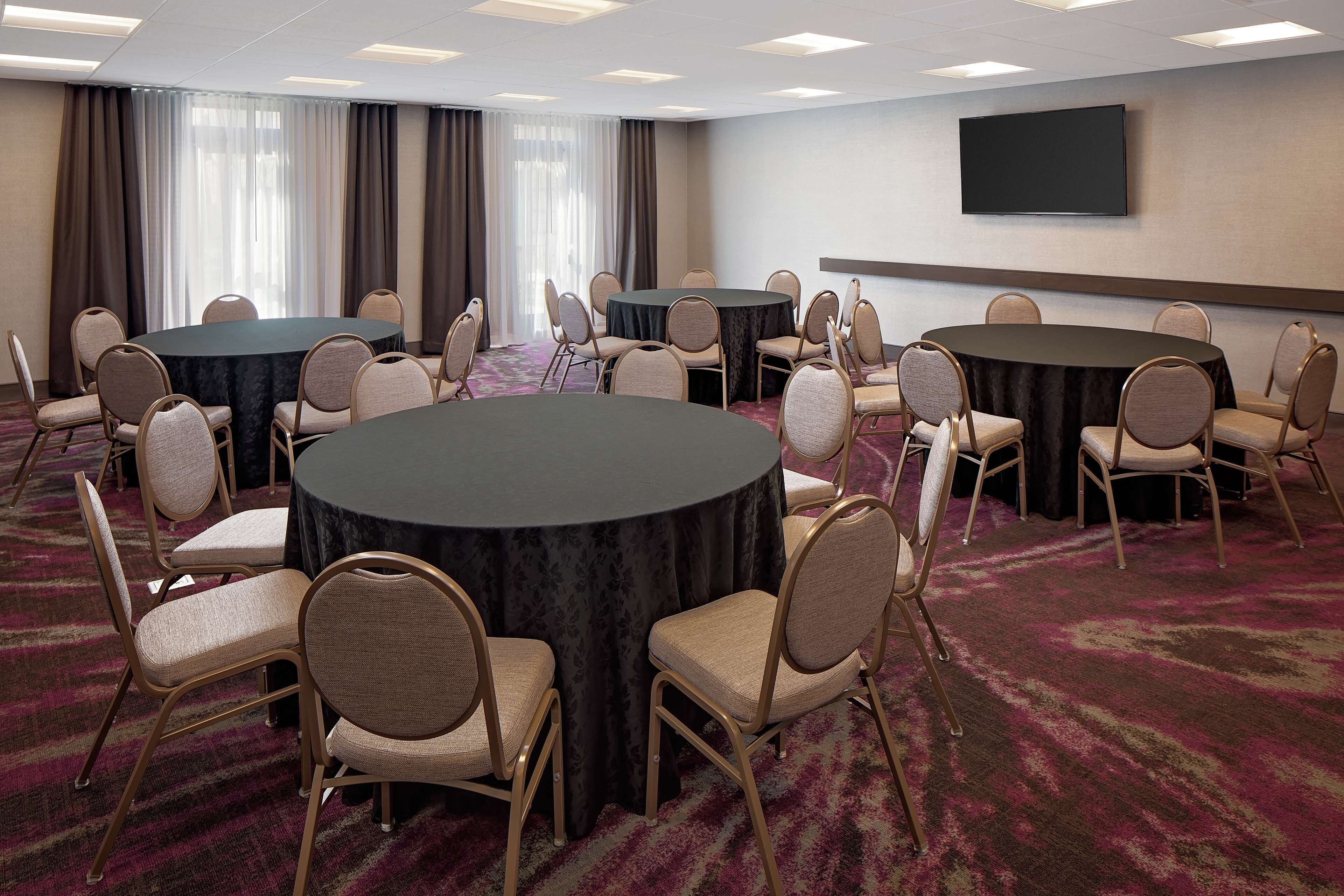Une salle de conférence équipée de tables et de chaises rondes, d’un téléviseur à écran plat et d’une grande fenêtre avec des rideaux.