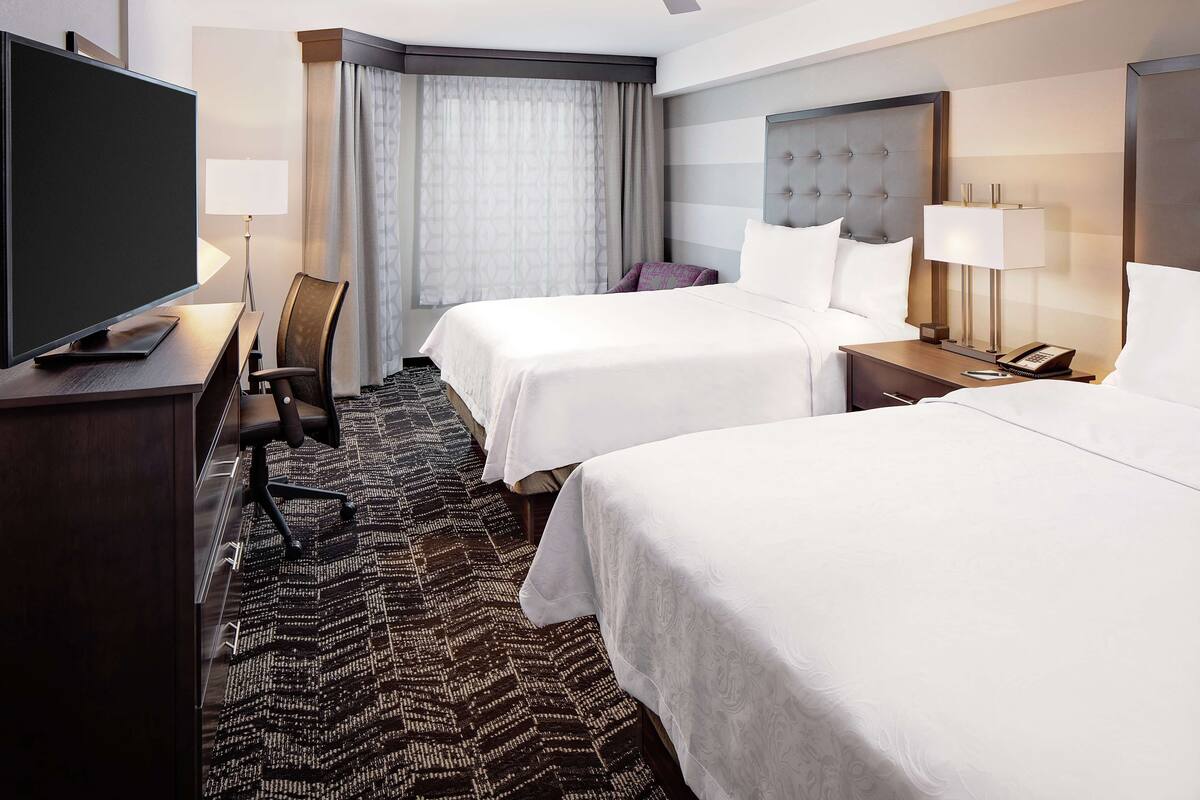 Ein Hotelzimmer mit zwei Betten, einem Schreibtisch mit Fernseher, einem Sessel, einer Lampe und einem Telefon.