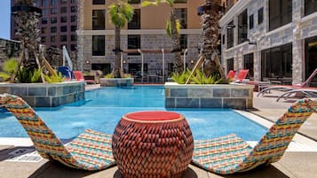 Un área de piscina con sillones de descanso y una mesa redonda, rodeada de palmeras y un edificio de piedra.