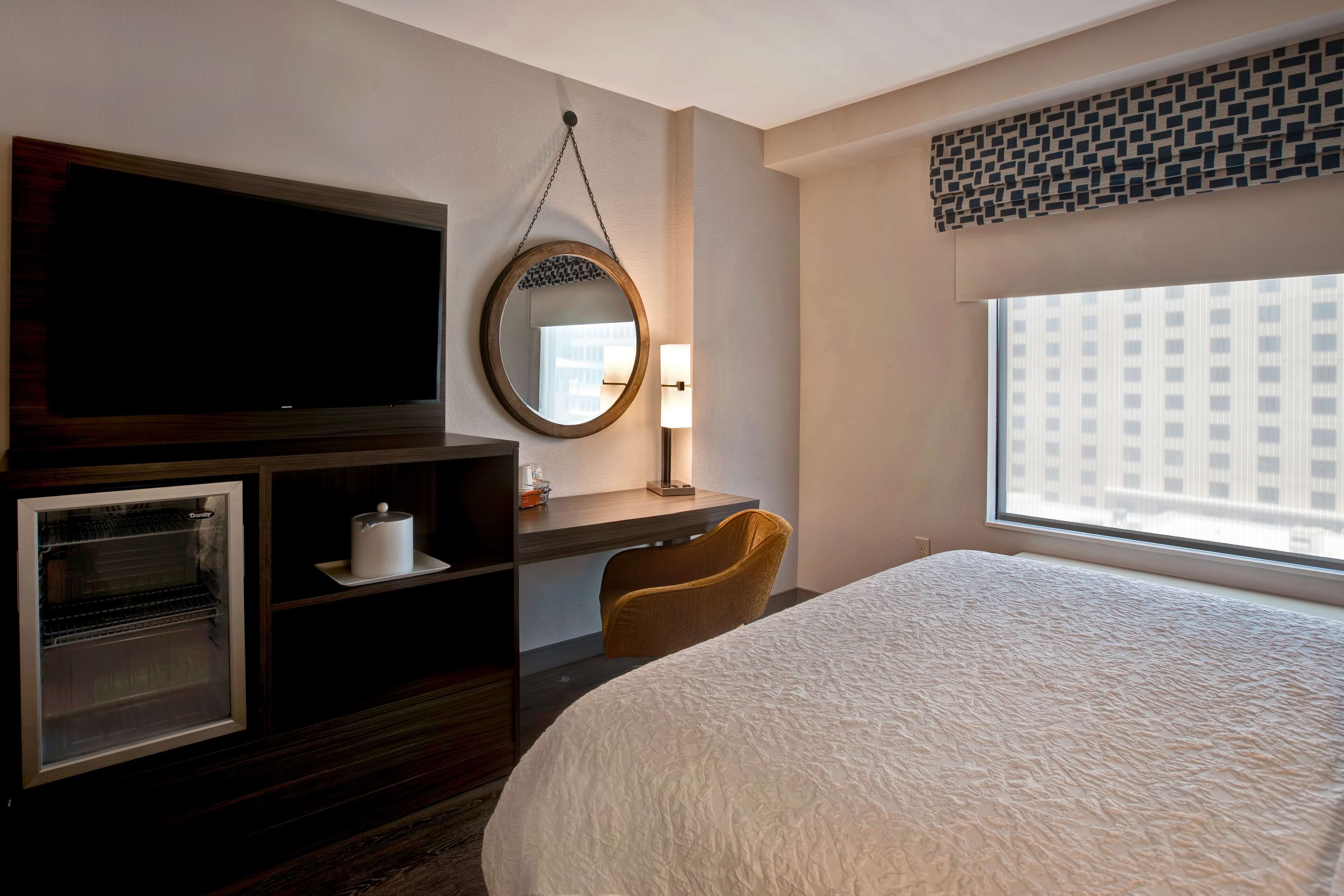 Une chambre d’hôtel dotée d’un grand lit, d’une télévision à écran plat, d’un minibar, d’un bureau avec une lampe et d’une fenêtre donnant sur des immeubles.