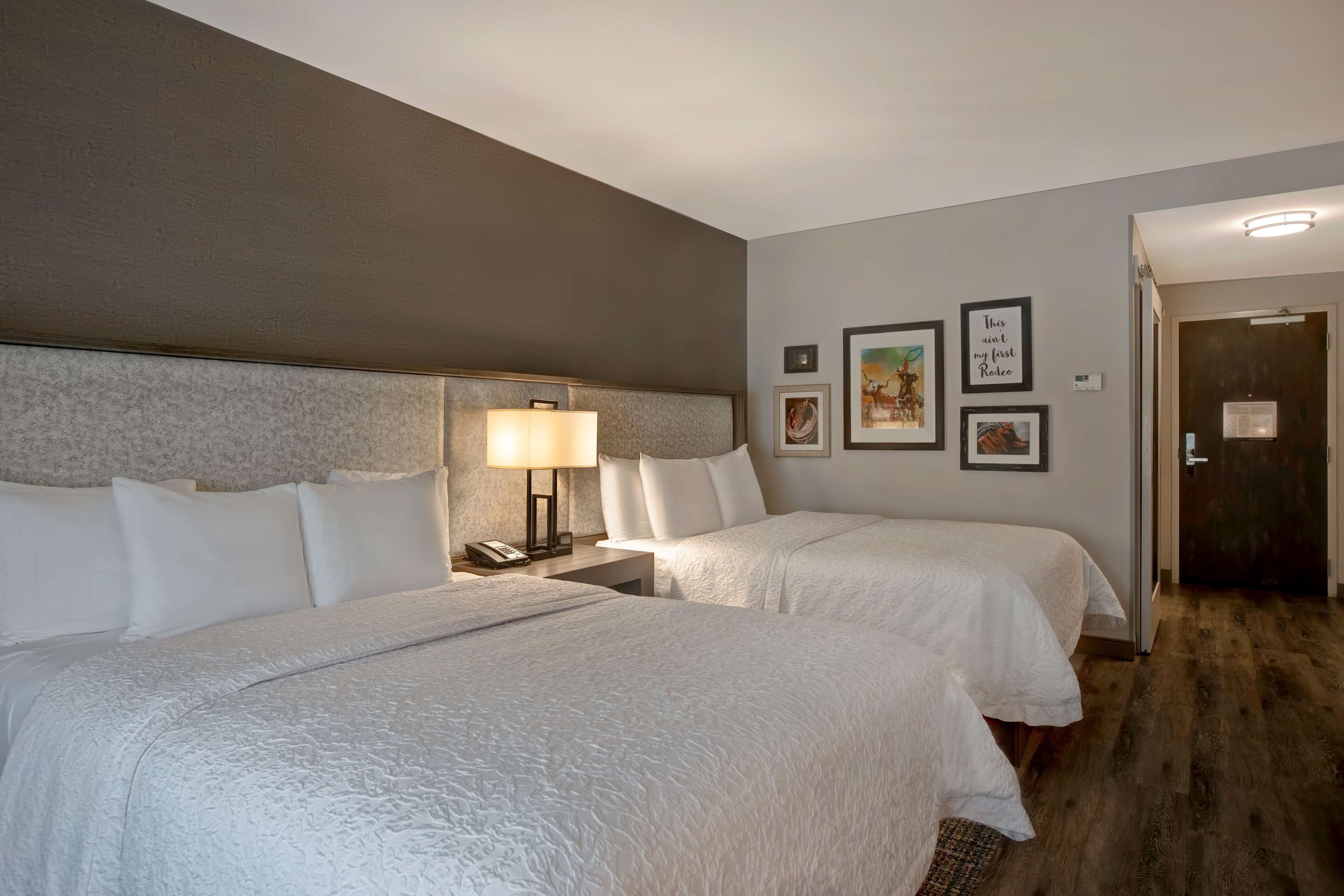 A hotel room with two beds, a nightstand with a lamp, and a door leading to another room.