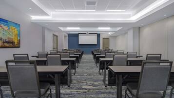 A conference room with rows of tables and chairs, a projector screen, and a carpeted floor.