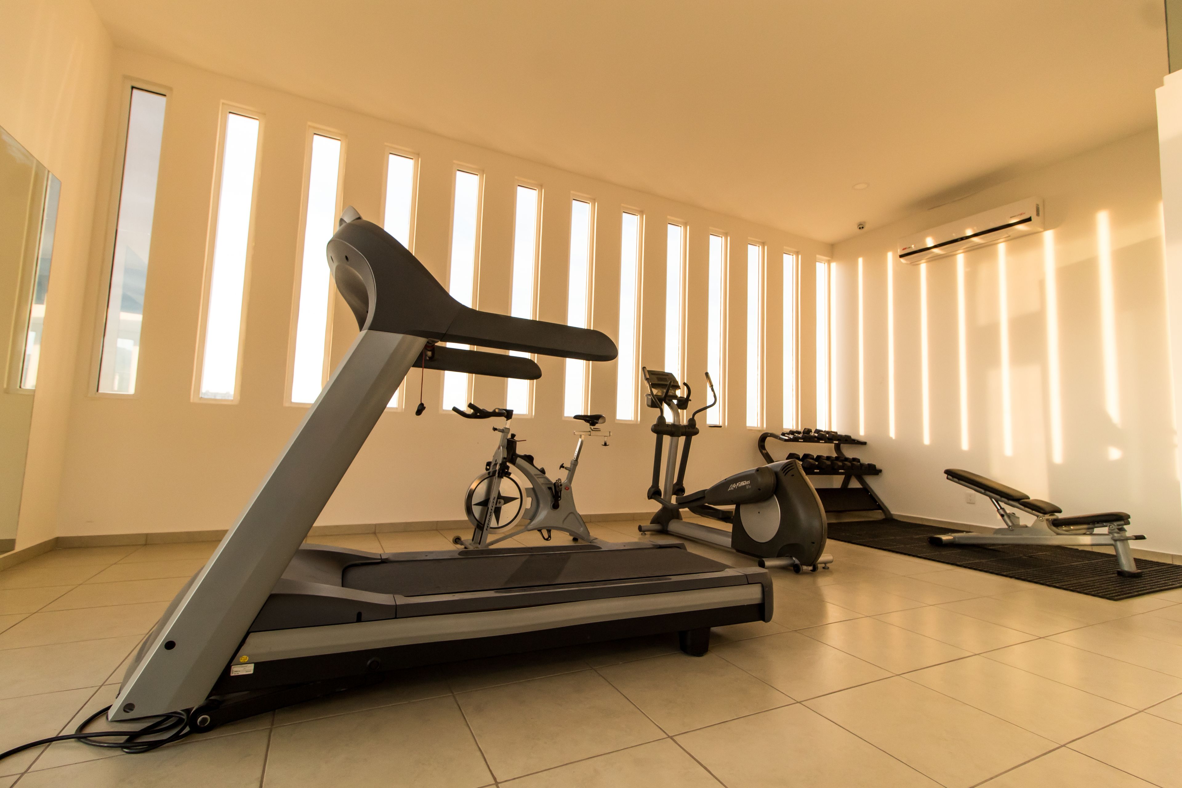 Uma academia com esteira, bicicletas ergométricas e um banco.