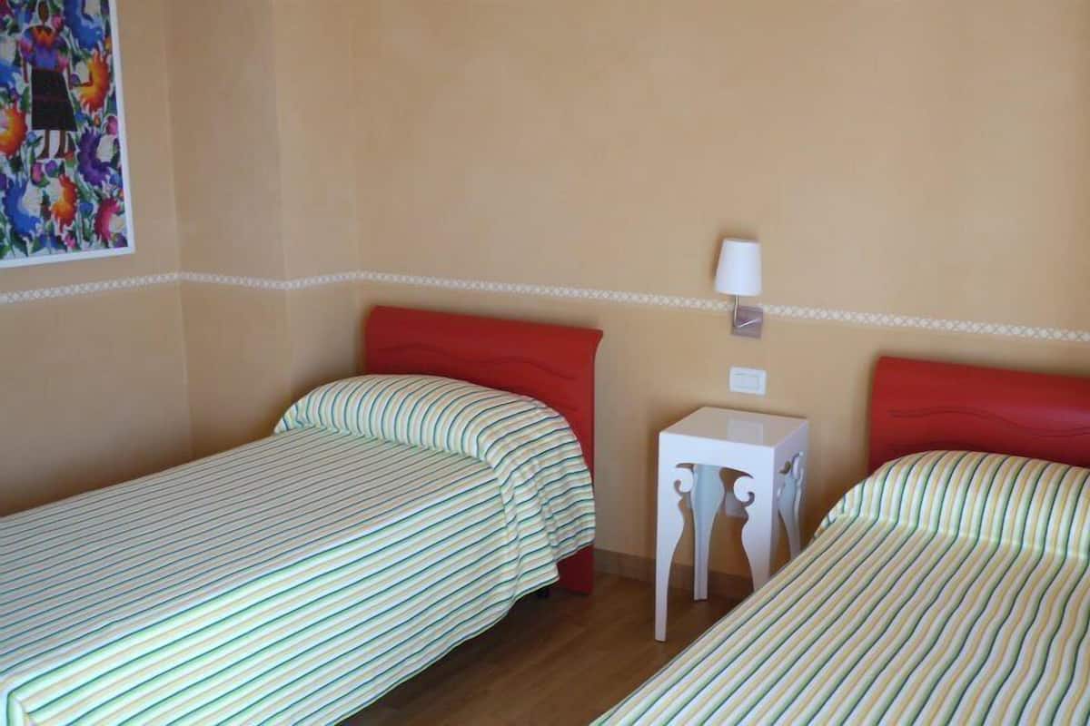 A room with two single beds, each with striped bedding and red headboards. There is a small white nightstand with a lamp between the beds.