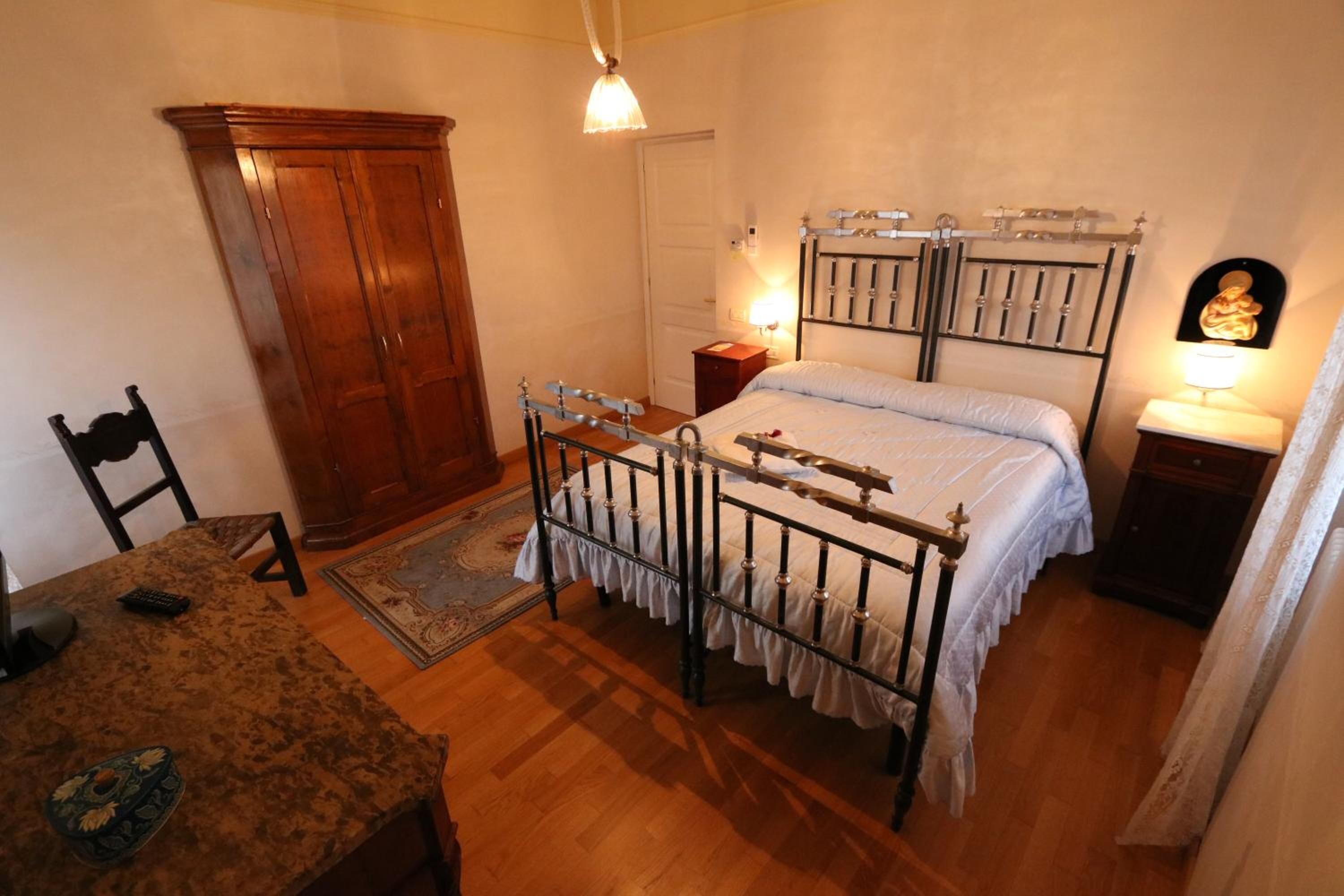 Un dormitorio con un armario de madera, una cama con estructura metálica, una mesita de noche con una lámpara y una pequeña mesa con un cuenco decorativo.