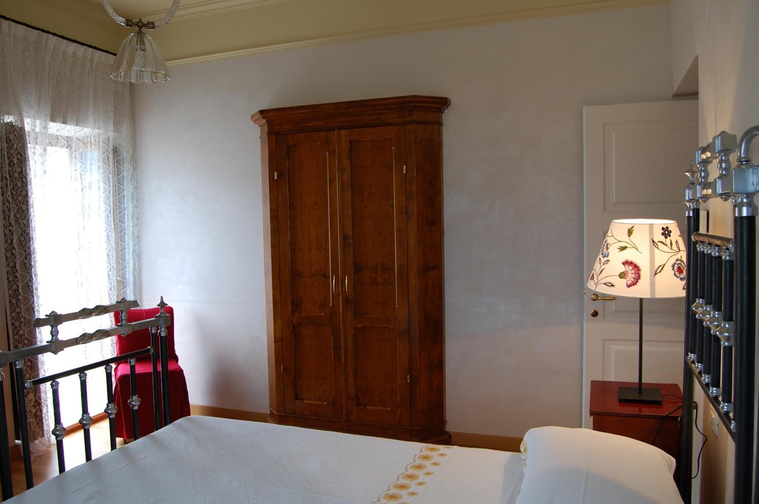 A bedroom with a wooden wardrobe, a bed with white bedding, a nightstand with a lamp, and a window with sheer curtains.