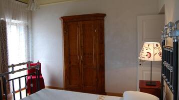 A bedroom with a wooden wardrobe, a bed with white bedding, a nightstand with a lamp, and a window with sheer curtains.