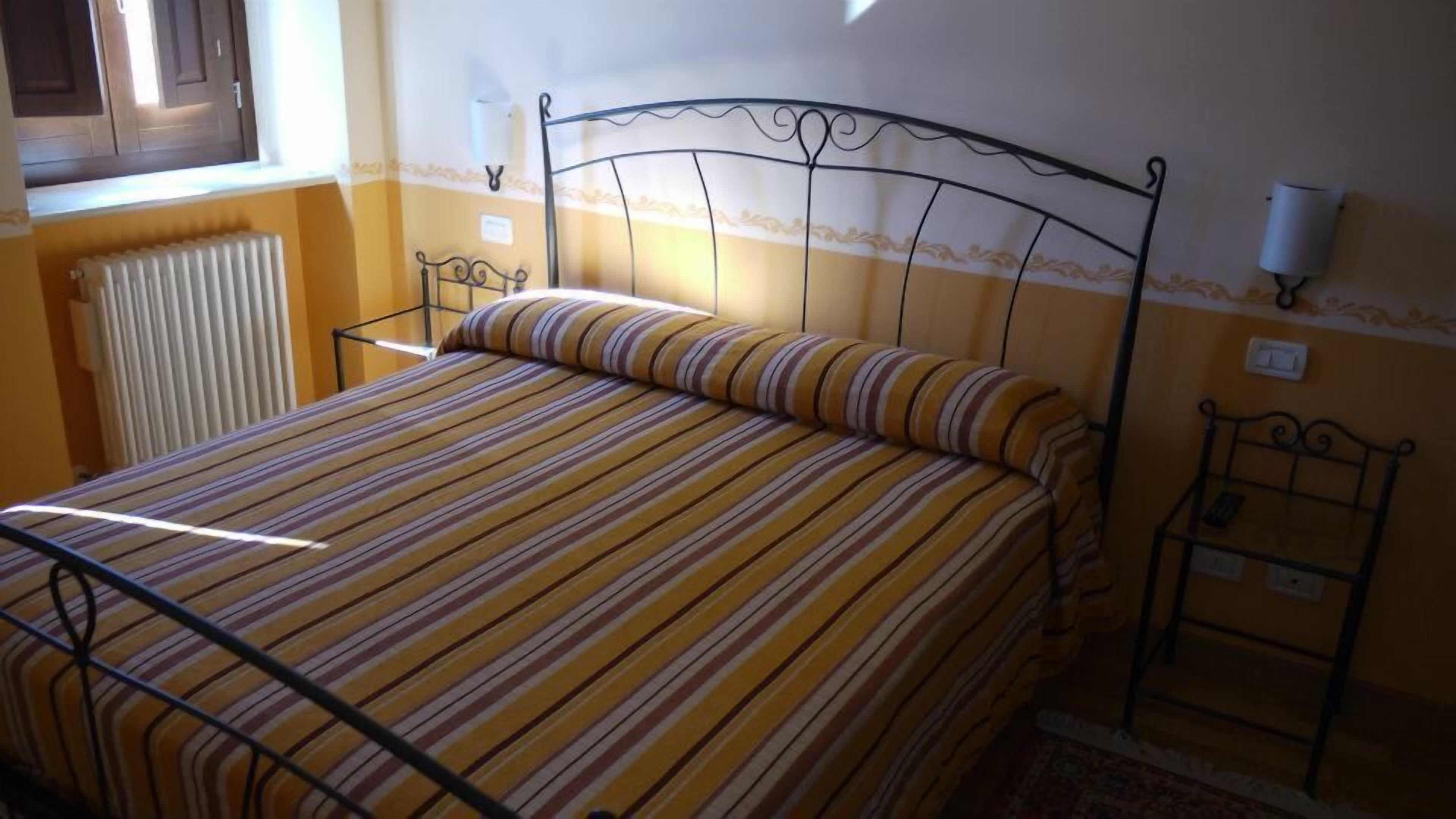 Cama com colcha listrada, radiador, luminária na parede e janela com persianas.