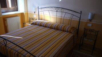 Cama com colcha listrada, radiador, luminária na parede e janela com persianas.