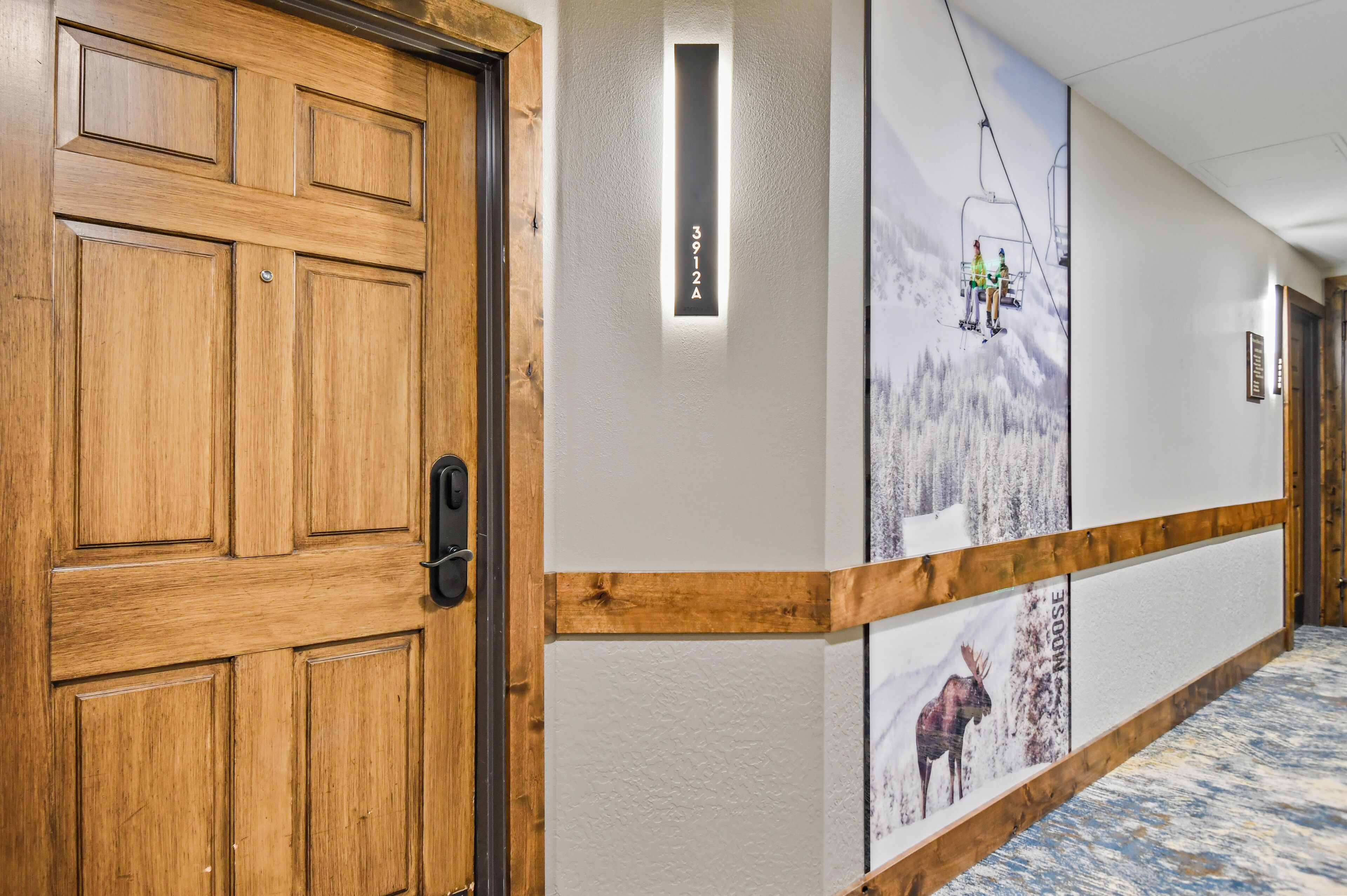A hallway with a wooden door, a wall-mounted sign with the number 3912A, and a ski lift image on the wall.