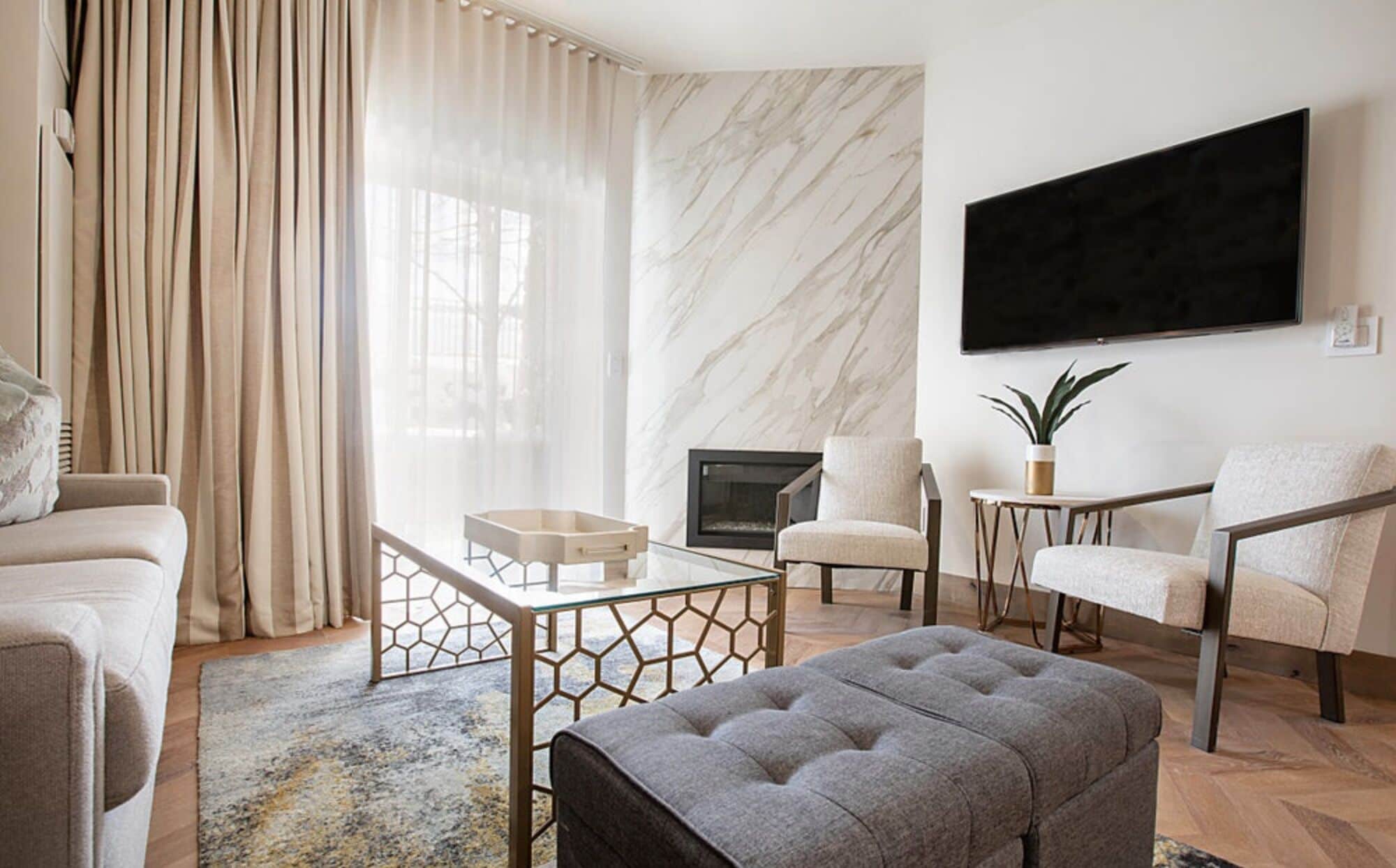Un salon moderne comprenant un canapé, un pouf et des fauteuils, une table basse en verre, une cheminée et un téléviseur fixé au mur.