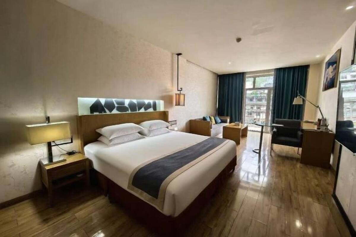 Habitación de hotel con una cama grande, un escritorio, una silla, una lámpara y vistas a la ciudad desde la ventana.