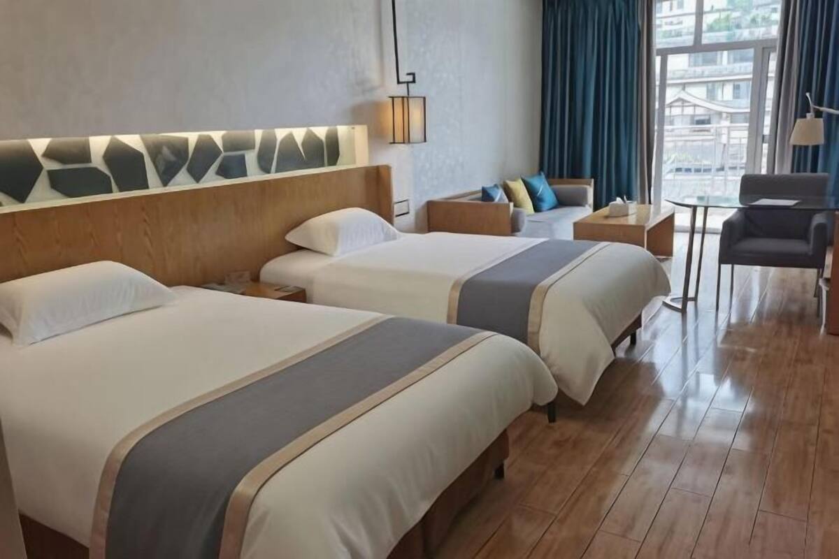 Habitación de hotel con dos camas, cabecero de madera, una mesita y vista al exterior.