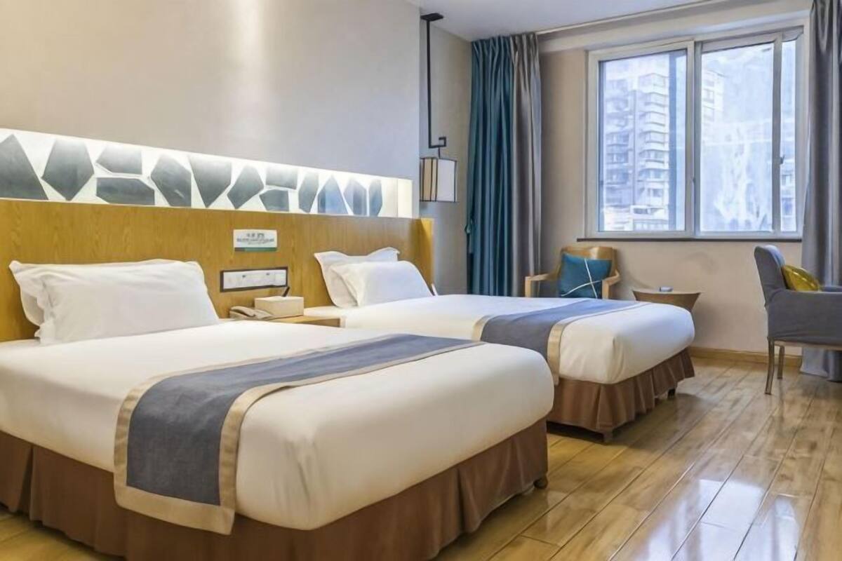 Habitación de hotel con dos camas, cabecero de madera, una mesita, una silla y un ventanal con cortinas.
