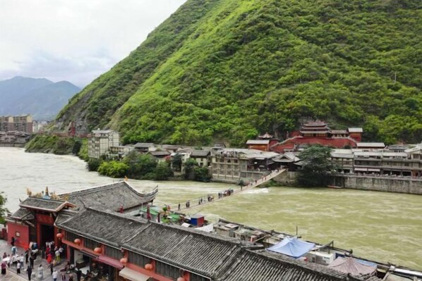 A traditional Chinese building with a steeply sloped roof, located beside a river with a bridge connecting to a hillside. - Huangqiao Hotel (Luding)
