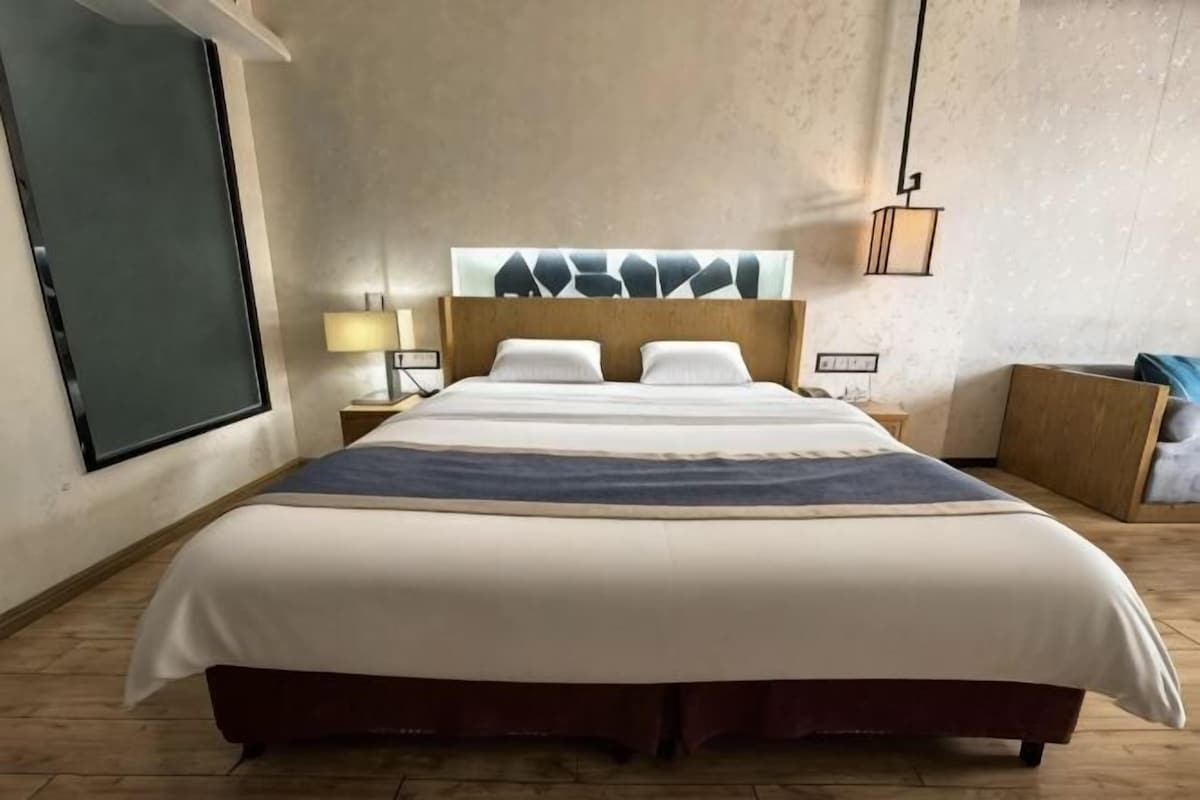 A hotel room with a large bed, a nightstand, a lamp, and a view of the outdoors.