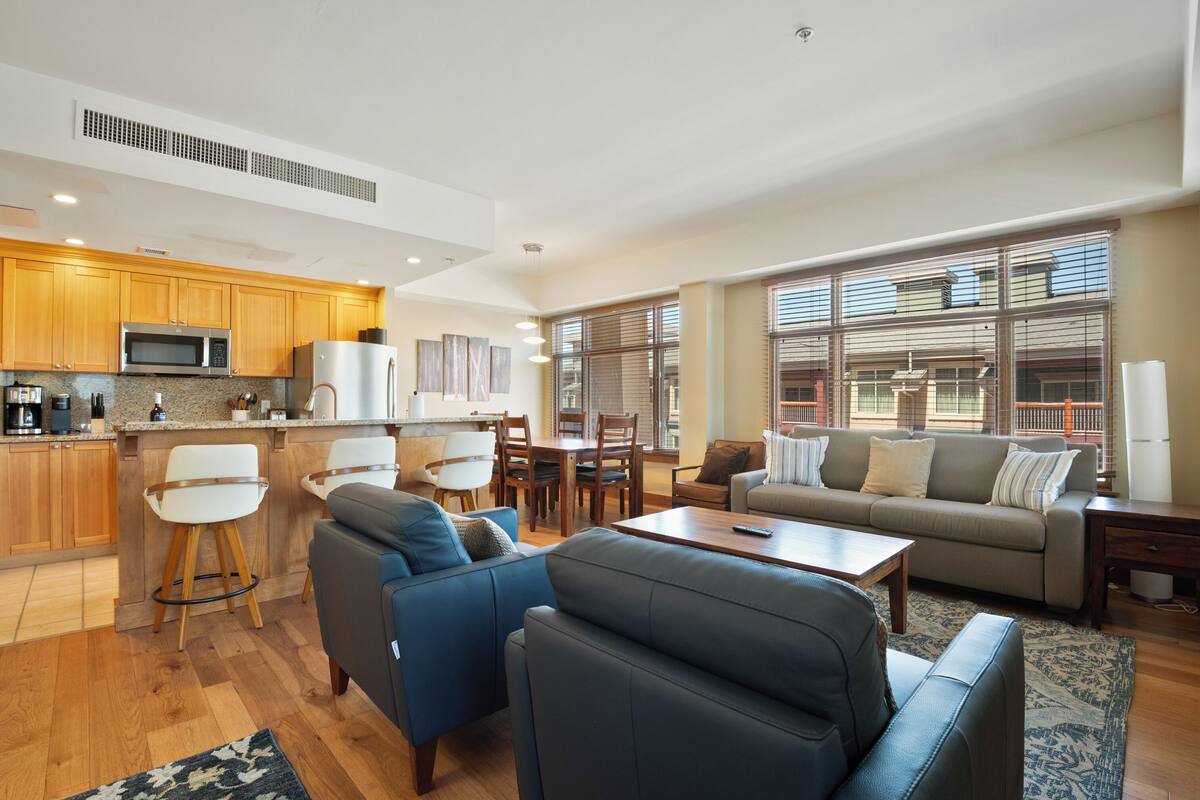 Eine moderne Küche mit Holzschränken, einer Mikrowelle und einem Kühlschrank. Es gibt einen Essbereich mit Tisch und Stühlen sowie ein Wohnzimmer mit Sofa und Couchtisch.