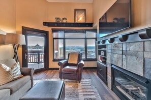 A living room with a fireplace, a leather armchair, a sofa, and a large window offering a view of the mountains. - Sundial Lodge (Park City)