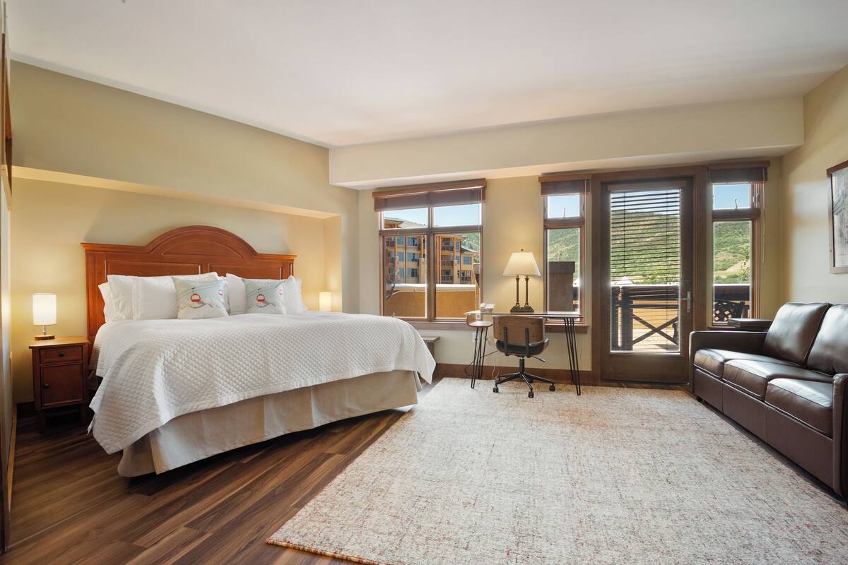 Ein Schlafzimmer mit einem großen Bett, einem Schreibtisch, einem Ledersofa und Blick auf ein Gebäude durch die Fenster.