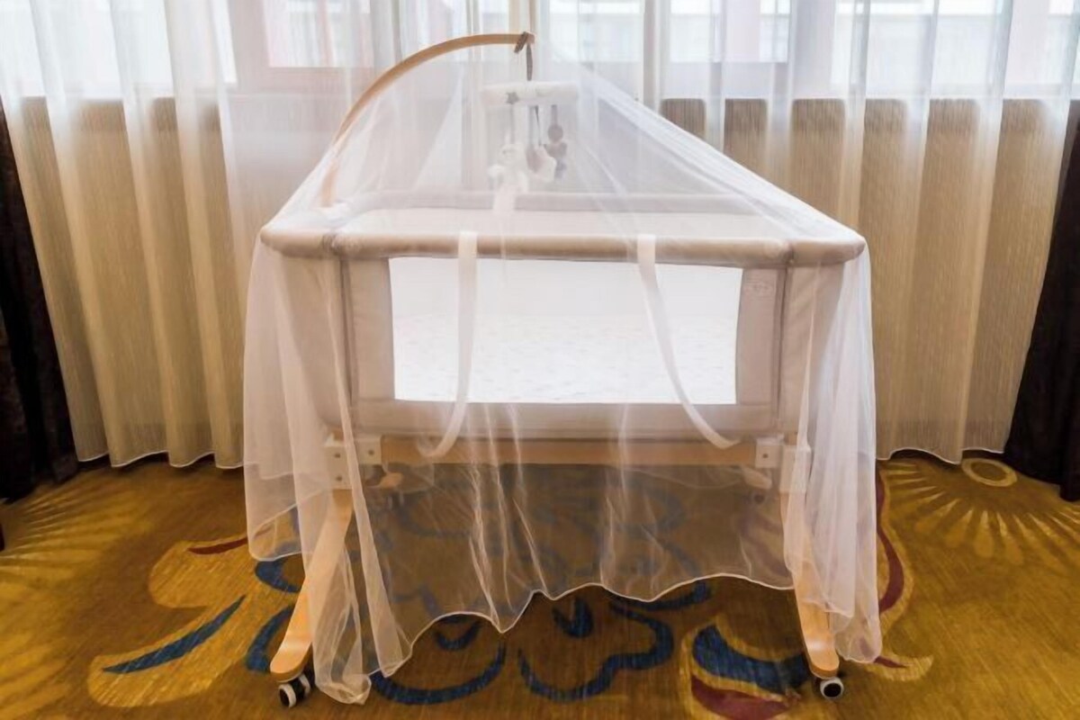 A crib with a white canopy in a room with a patterned carpet and sheer curtains.