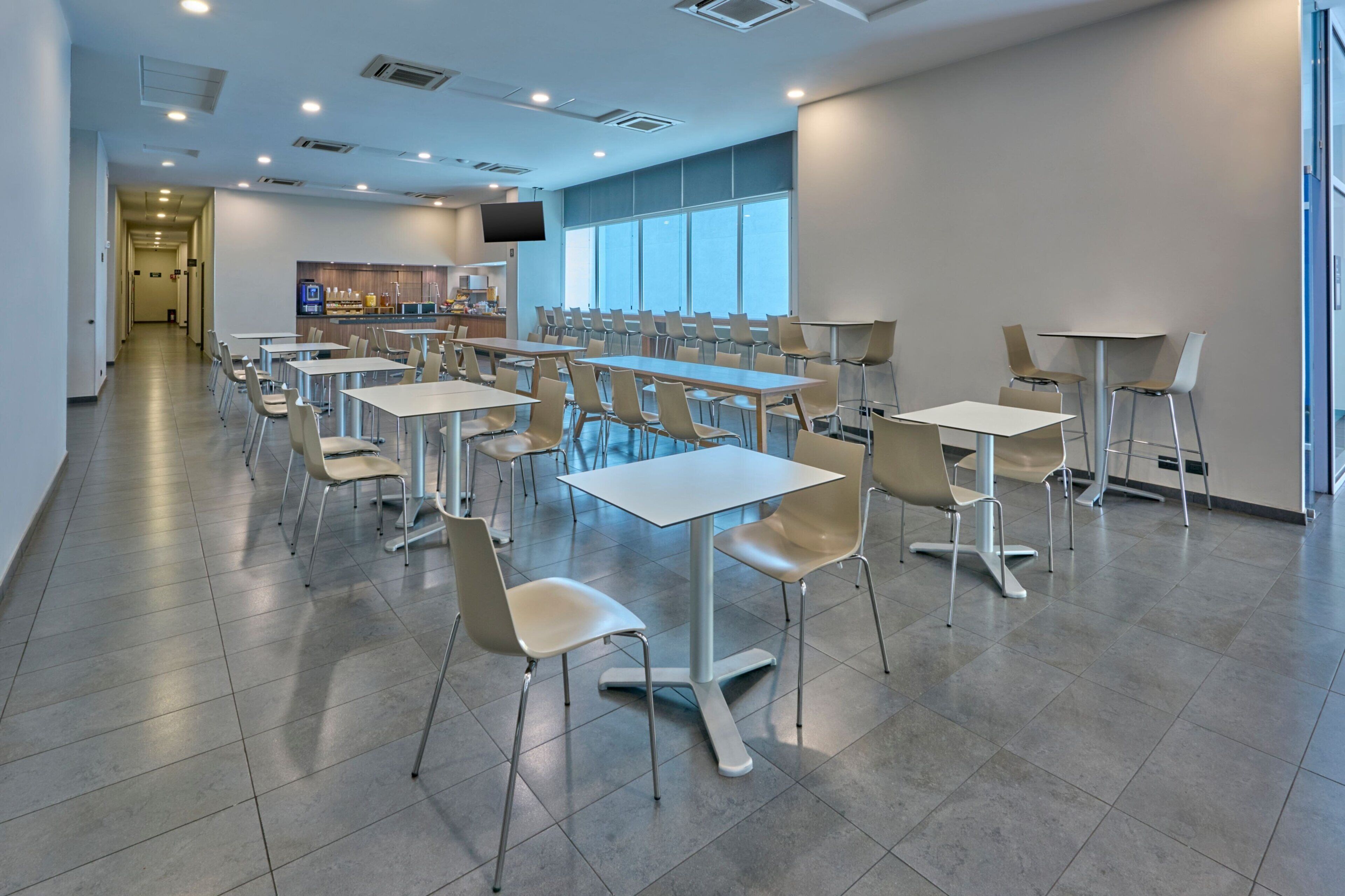 A modern dining area with multiple tables and chairs, a kitchen area with a TV, and large windows providing natural light.