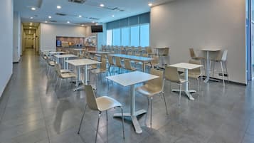 A modern dining area with multiple tables and chairs, a kitchen area with a TV, and large windows providing natural light.