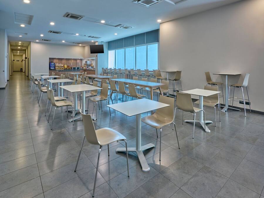 A modern dining area with multiple tables and chairs, a kitchen area with a TV, and large windows providing natural light.