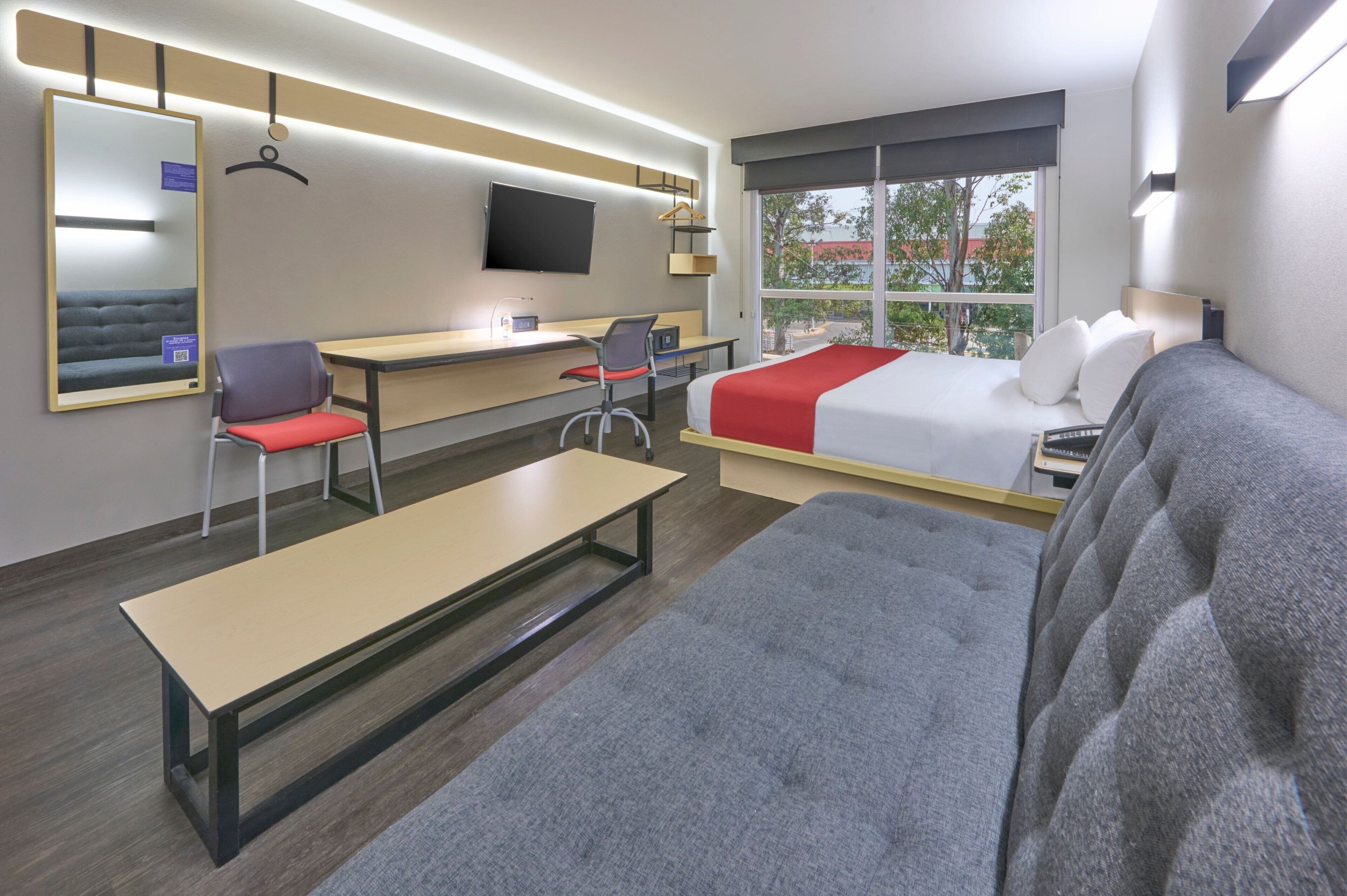 A modern hotel room with a bed, a desk, a chair, a bench, a TV, and a large window.