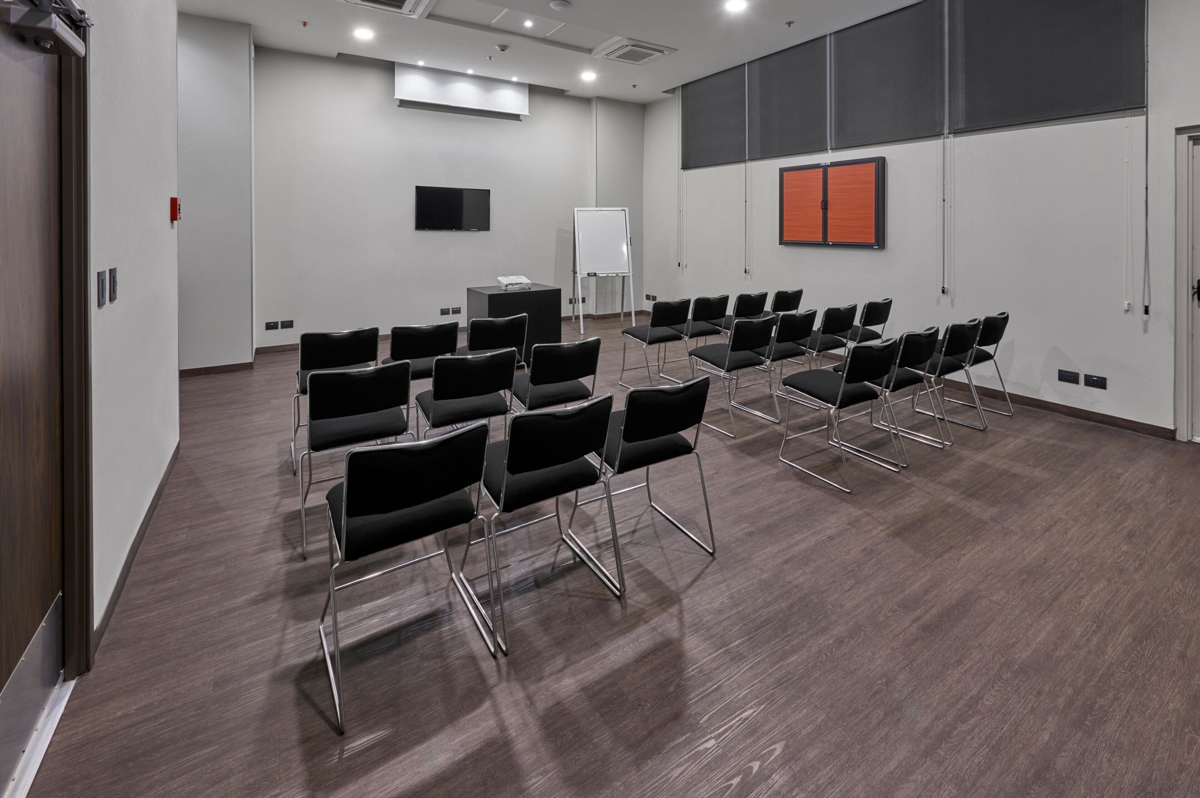 Un salón de conferencias con filas de sillas negras, una pizarra, una pantalla de proyector y un televisor de pantalla plana en la pared.