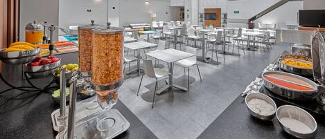 A modern hotel dining area with buffet-style food stations, including fruits, cereals, and other breakfast items.