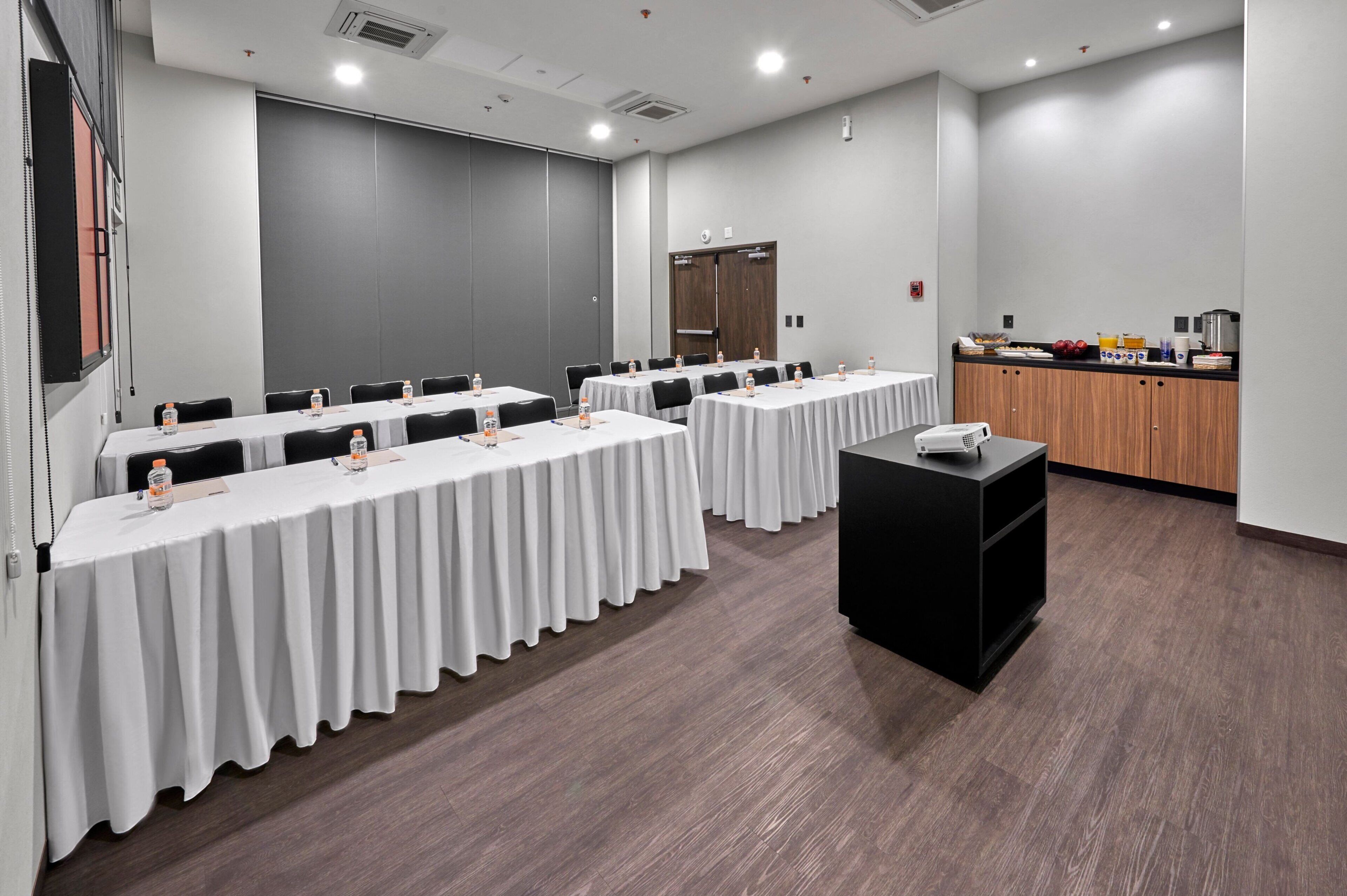 A conference room with multiple tables, chairs, a podium, and a kitchenette area.