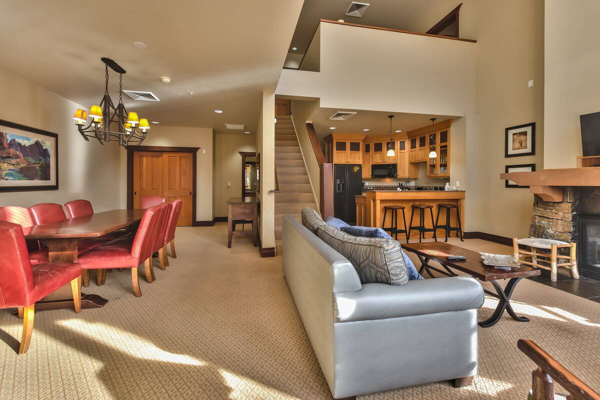 A spacious living room with a high ceiling, a dining area with red chairs, a light blue sofa, and a kitchen with wooden cabinets and a stone fireplace.