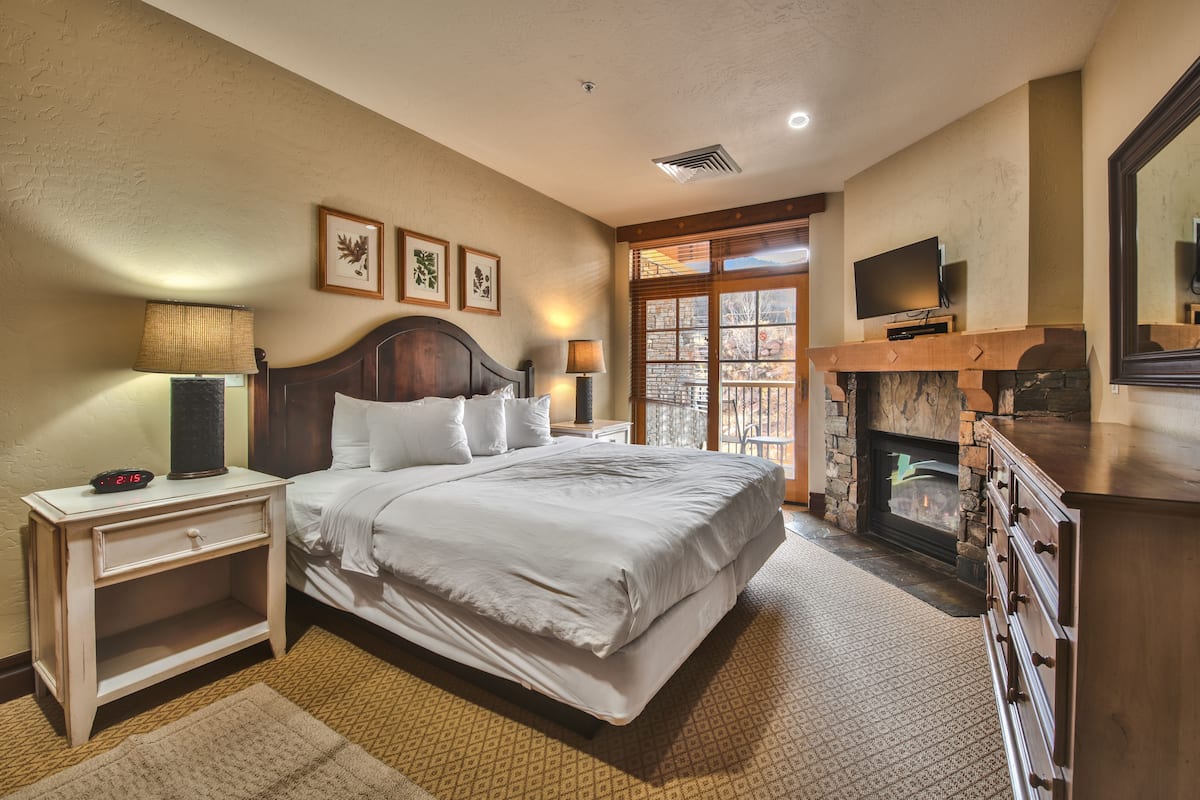 A bedroom with a large bed, a nightstand, a dresser, a fireplace, and a TV.