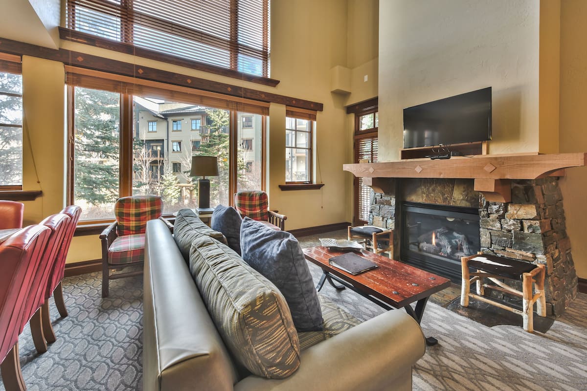 A spacious living room with a stone fireplace, a large flat-screen TV mounted above it, and a comfortable seating area with a sofa and chairs.