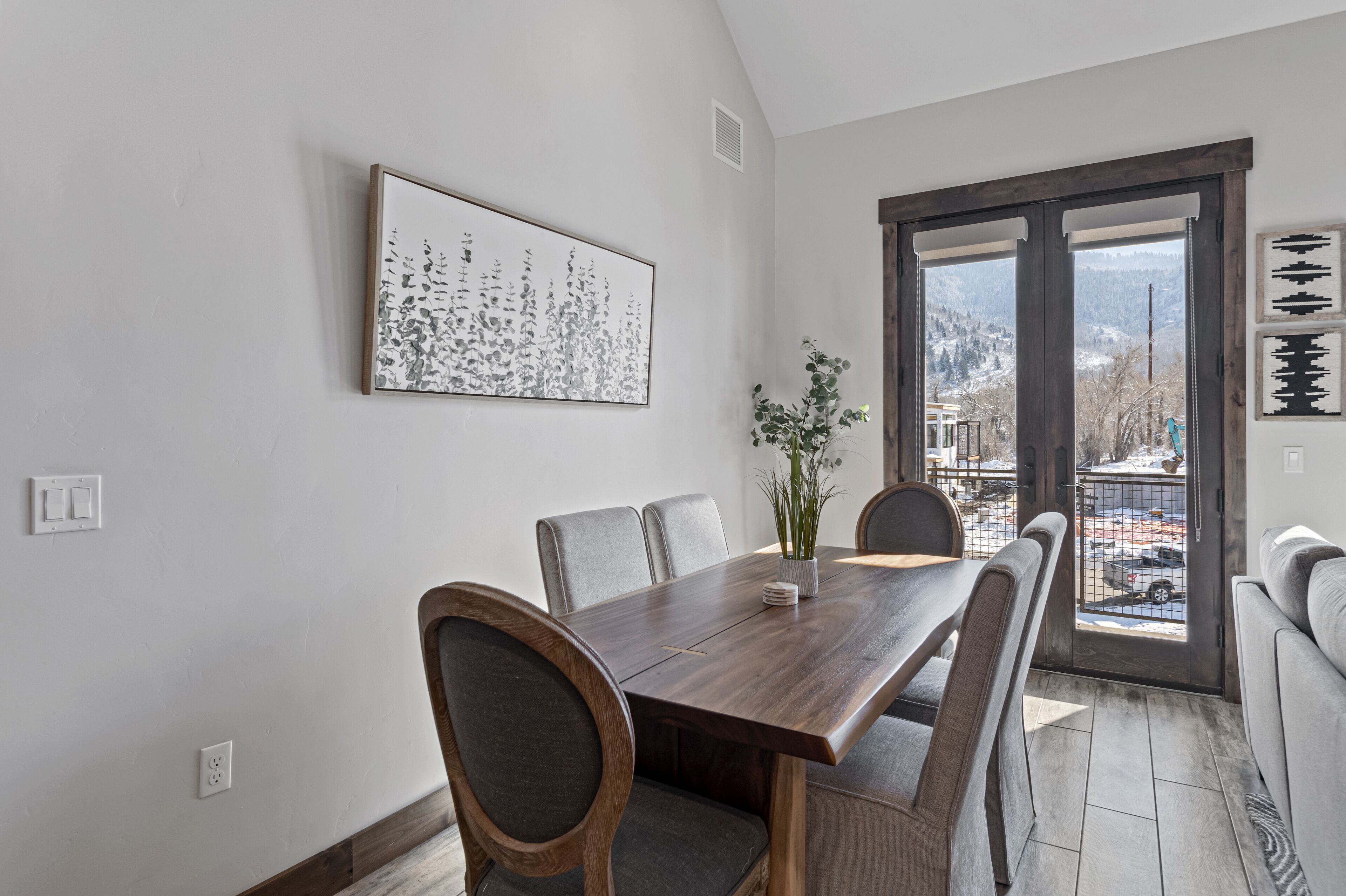 A dining area with a wooden table and chairs, a potted plant, and a view through a glass door.