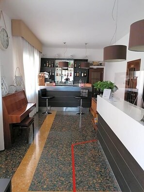 A hotel lobby with a reception desk, a bar area, a seating area with a bench, and a clock on the wall.