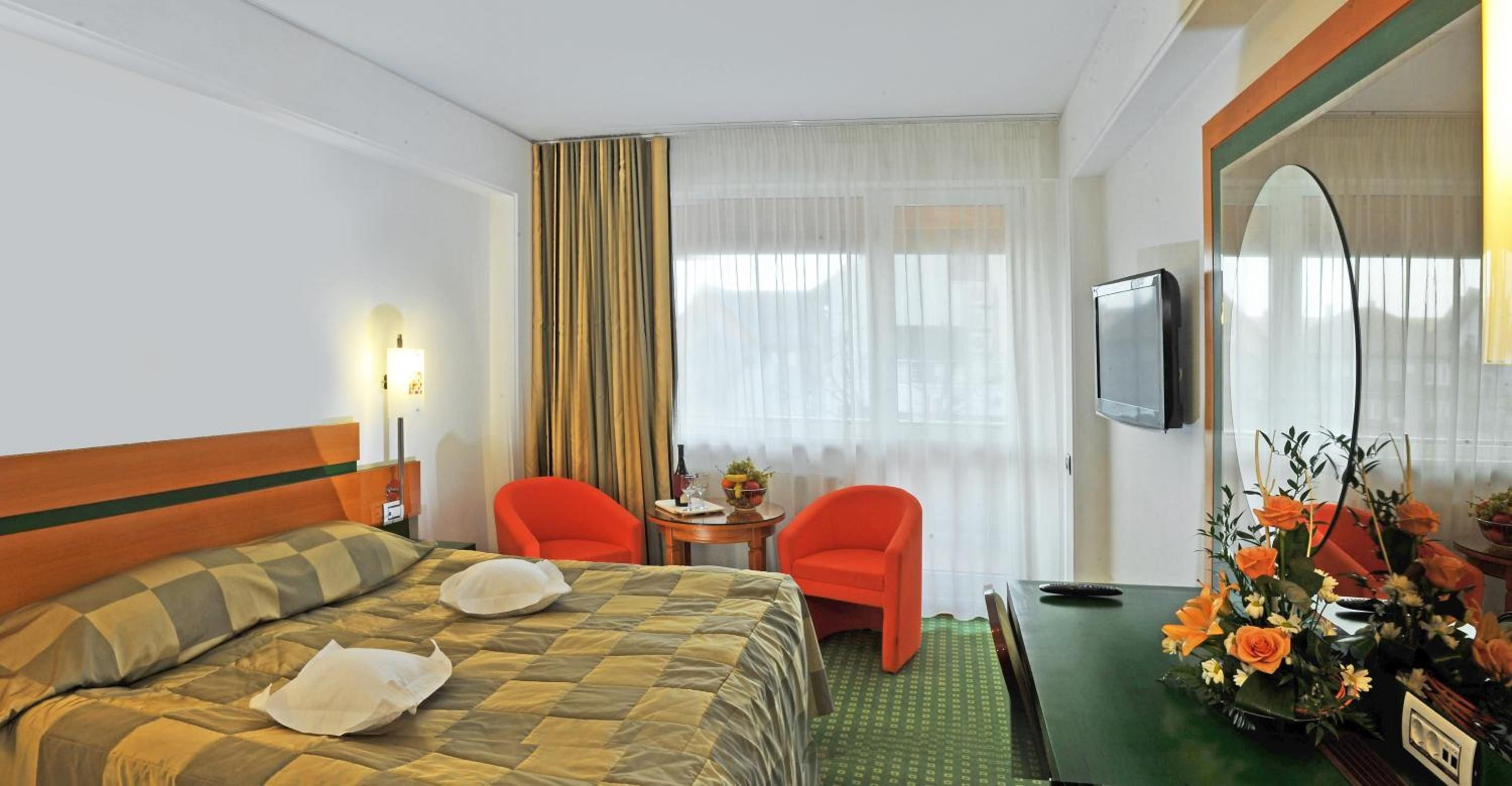 A hotel room with a bed, two chairs, a small table with a vase of flowers, a television, and a window with curtains.
