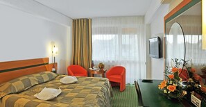 A hotel room with a bed, two chairs, a small table with a vase of flowers, a television, and a window with curtains.