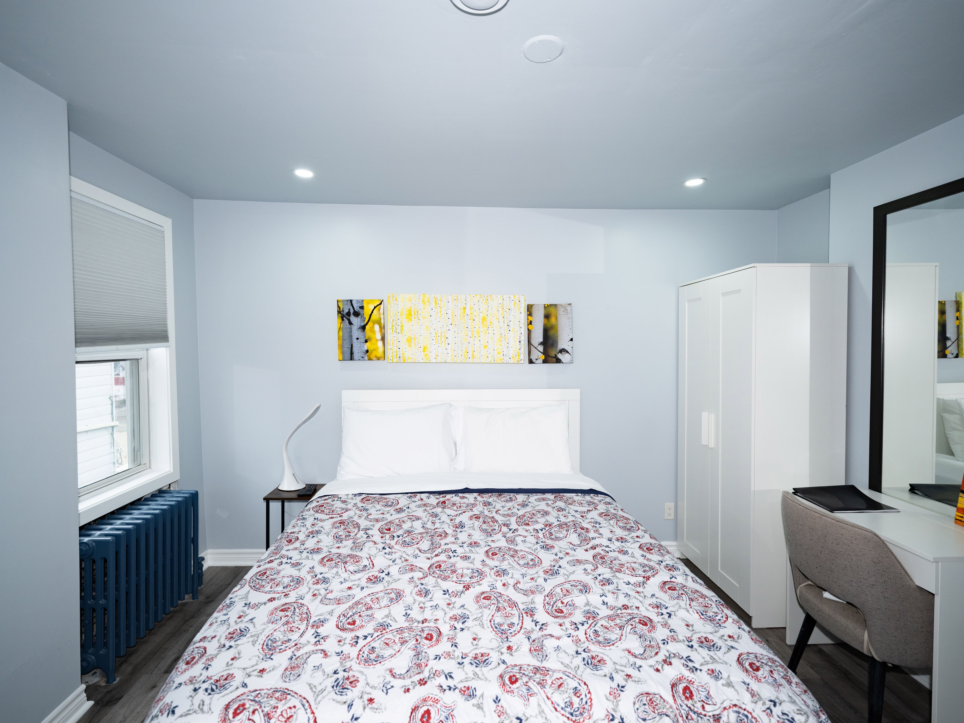 Une chambre à coucher comprenant un lit, un bureau, une chaise, un radiateur, une fenêtre avec des stores et des œuvres d’art accrochées au mur.